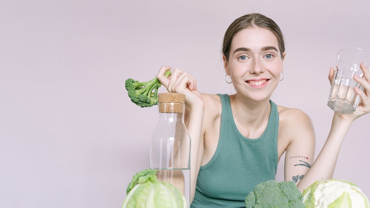 Woman in tank top smiling with broccoli, cabbage, and glass of water on table.